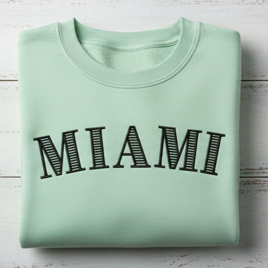 Folded mint green sweatshirt with 'MIAMI' print on a light wooden surface using striped serif font